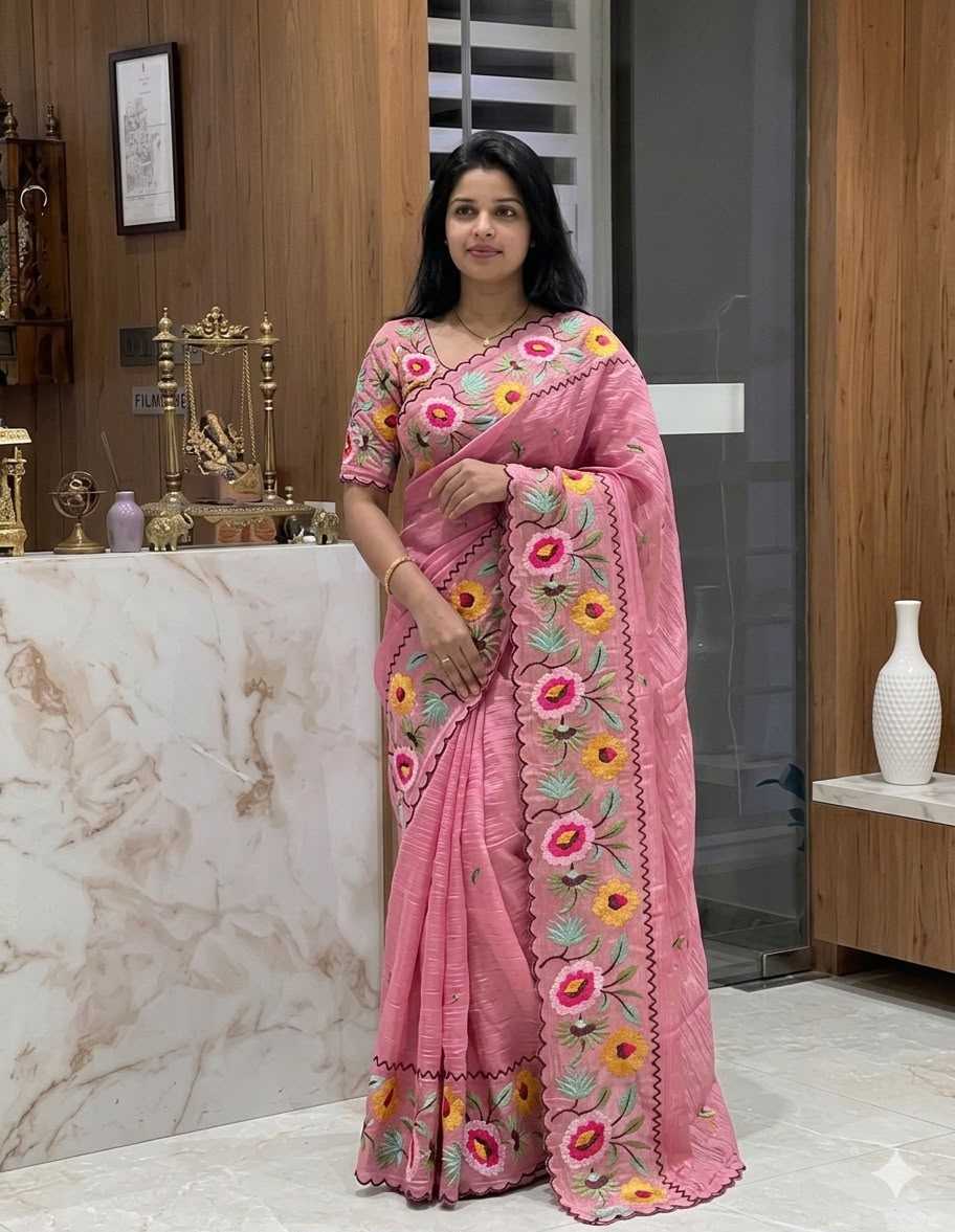 A woman wearing a pink embroidered saree with floral patterns, standing in an indoor setting with wood-paneled walls and a marble counter.