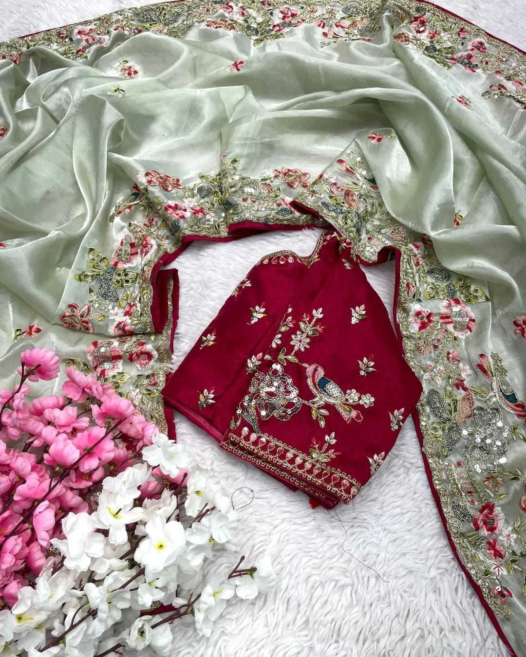 Embroidered red blouse with floral patterns on a white saree with floral border, surrounded by flowers.