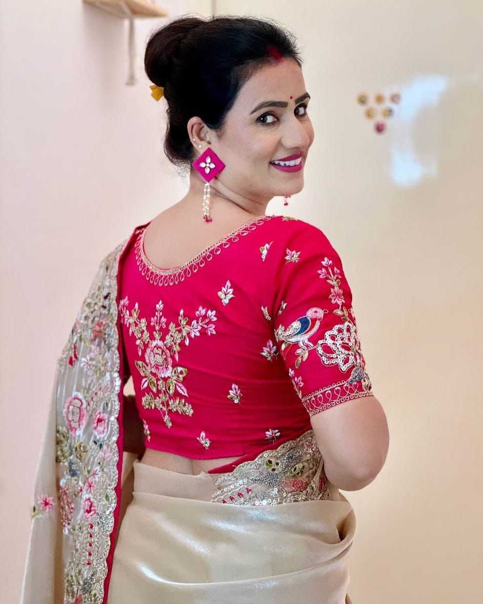Woman wearing a red blouse with floral patterns and a beige saree, smiling against a light background.