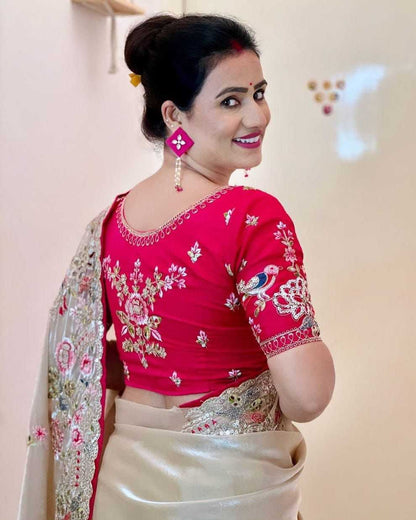 Woman wearing a red blouse with floral patterns and a beige saree, smiling against a light background.