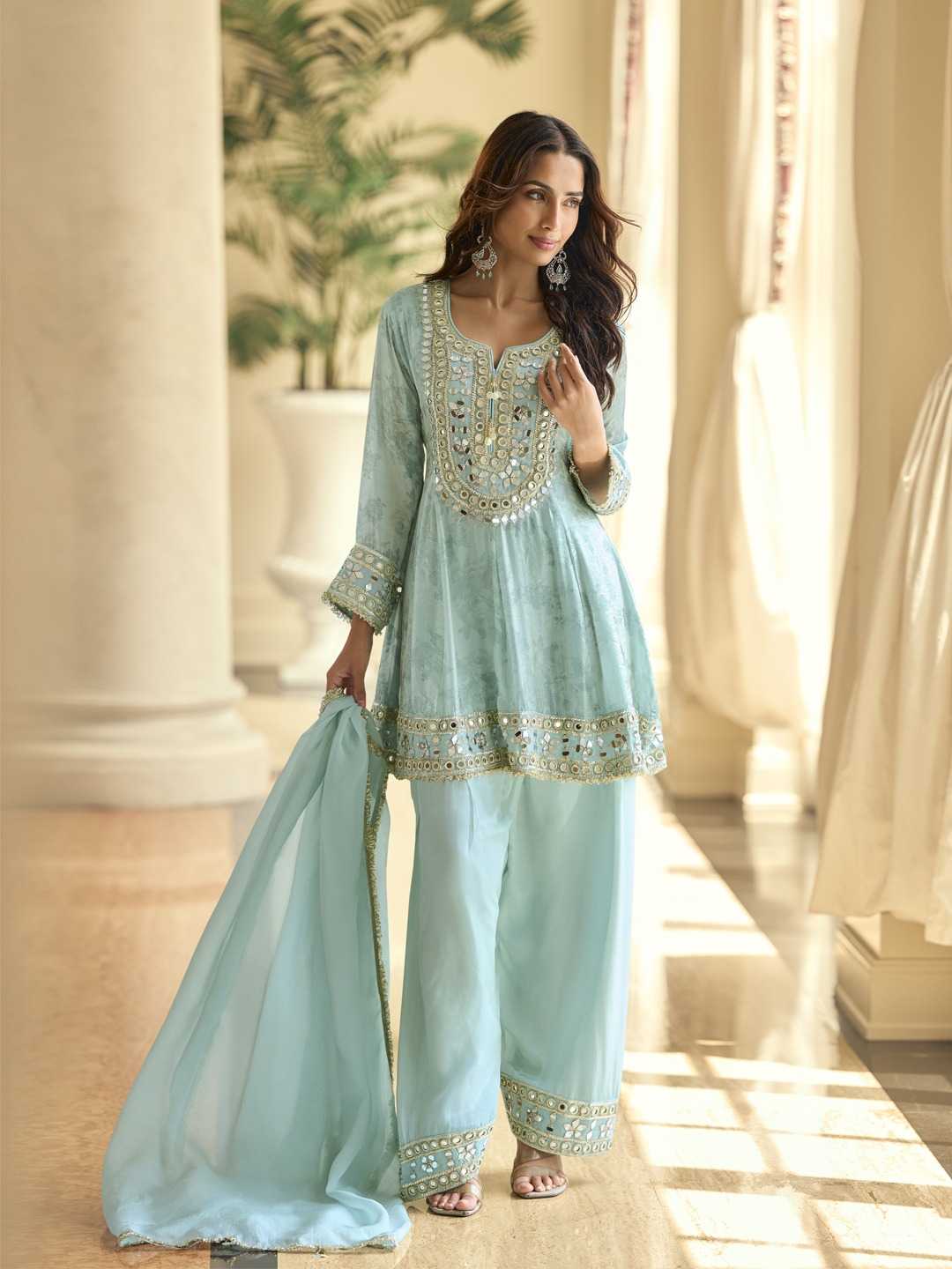 Woman in a light blue traditional outfit with gold embroidery standing in a sunlit room.