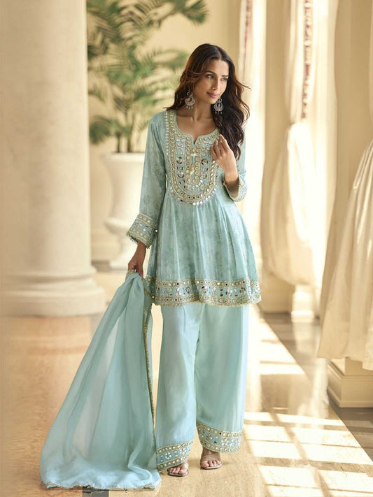 Woman in a light blue traditional outfit with gold embroidery standing in a sunlit room.