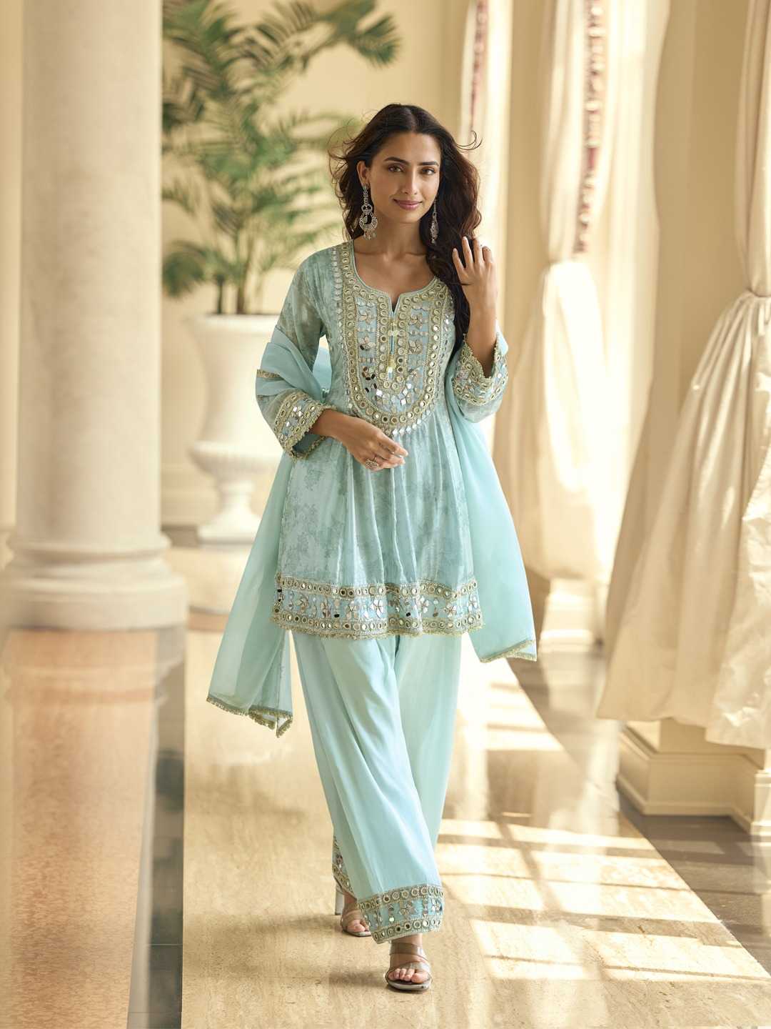 Woman in a light blue traditional outfit with intricate designs in a sunlit room.