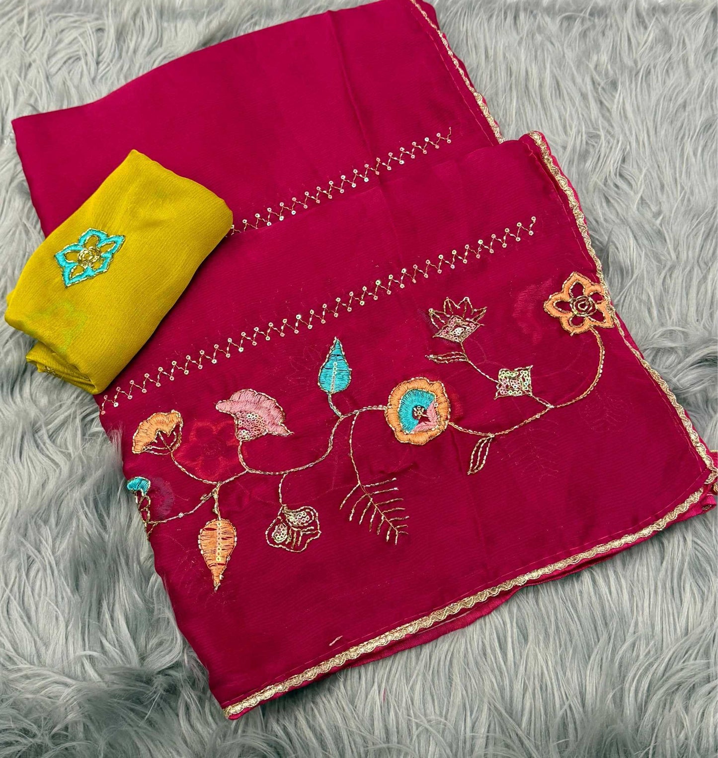 A folded magenta pink fabric piece with colorful floral embroidery and gold border trim, shown alongside a folded yellow fabric piece on a gray faux fur background.