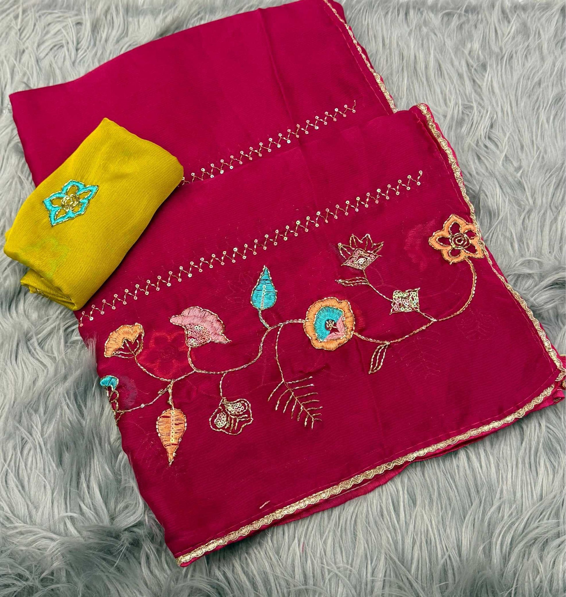 A folded magenta pink fabric piece with colorful floral embroidery and gold border trim, shown alongside a folded yellow fabric piece on a gray faux fur background.