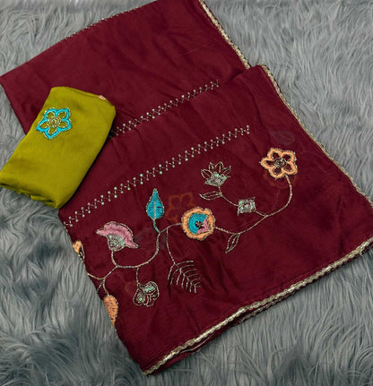 A folded deep maroon fabric set with colorful floral embroidery and a contrasting olive green fabric piece, displayed on a gray faux fur background.