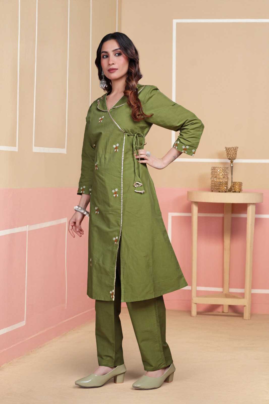 Woman wearing a green outfit with a long coat in a room with pink walls and a wooden table.