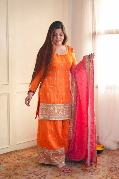 A woman modeling an orange traditional ethnic outfit with silver embroidery, holding a bright pink dupatta with gold borders in an indoor setting with cream walls and a patterned rug.