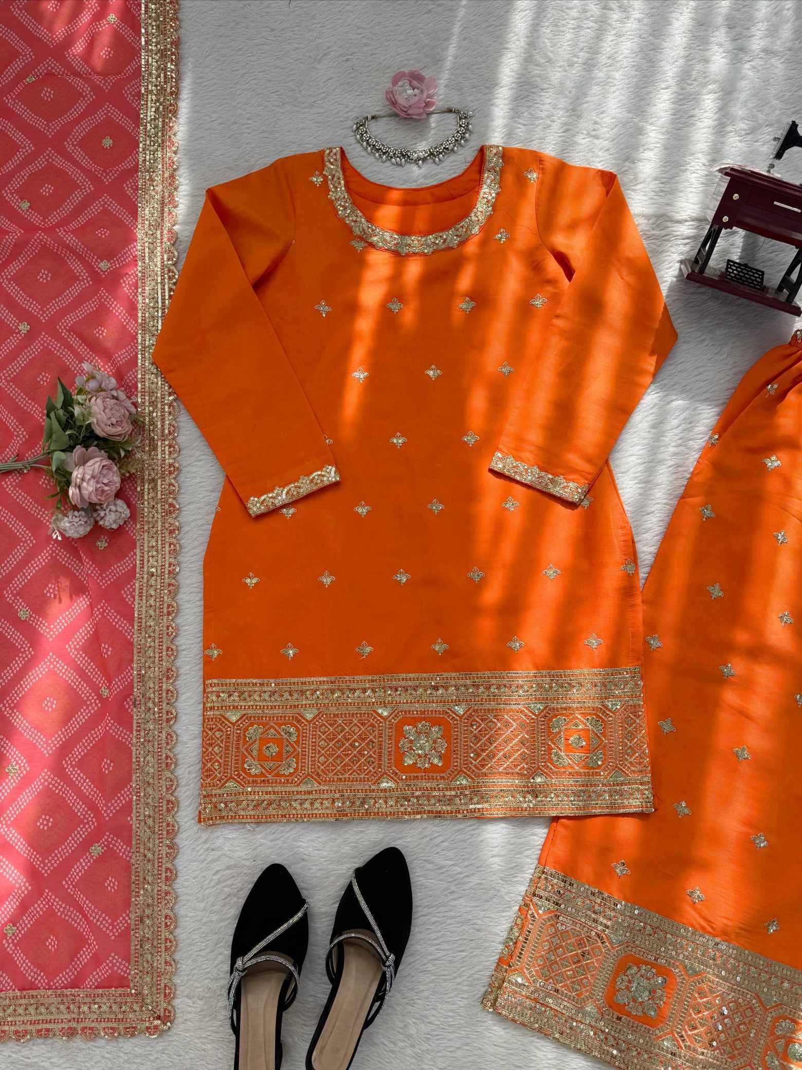 An orange traditional kurta set with gold embroidery, laid flat on a white textured surface with a pink dupatta, black flats, and decorative flowers.