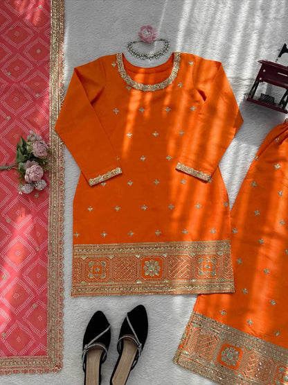 An orange traditional kurta set with gold embroidery, laid flat on a white textured surface with a pink dupatta, black flats, and decorative flowers.