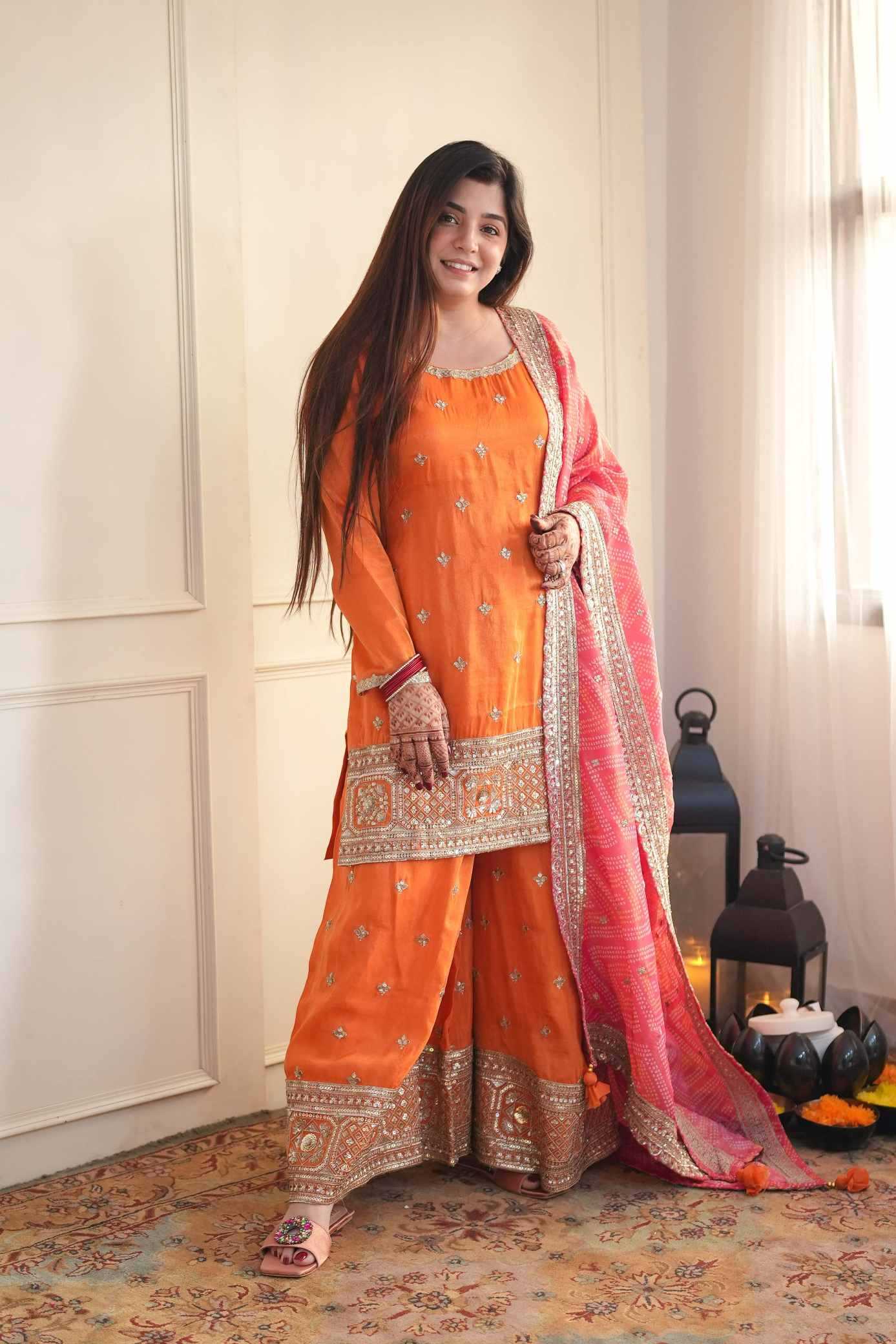 A woman modeling a traditional Indian outfit consisting of an orange kurta with silver embroidery, matching orange wide-leg pants, and a pink dupatta with gold borders, set against an indoor background with a patterned rug.