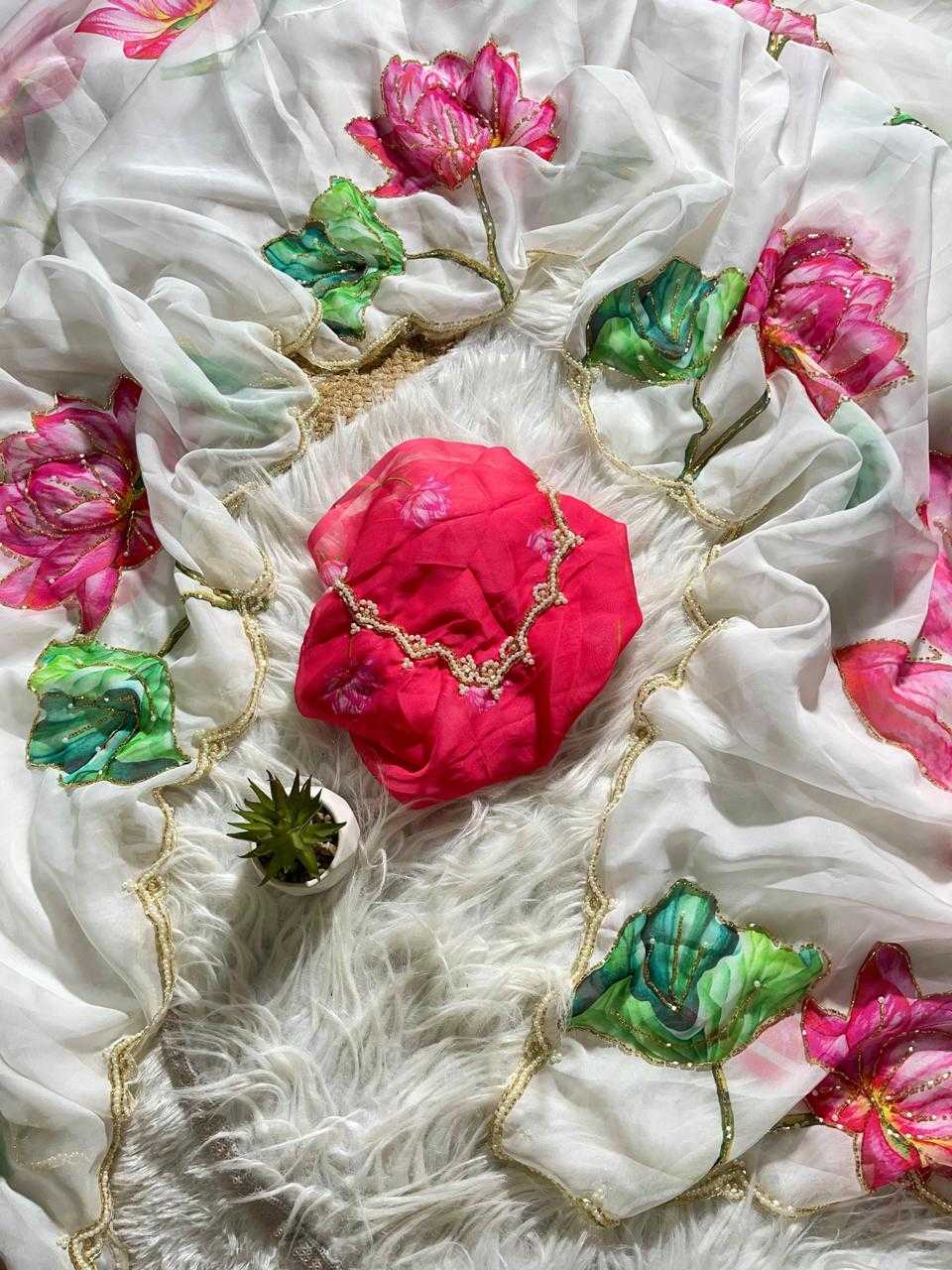 A white sheer fabric with pink and green floral prints and gold embroidered borders, styled on a white faux fur surface with a bright pink cloth and small green plant as props.