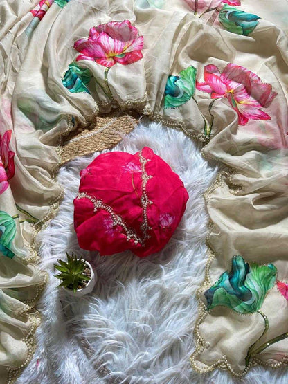 A beige printed scarf with pink and green floral motifs and gold border trim, arranged on a white faux fur surface with a bright pink wrapped headwrap and a small potted succulent.