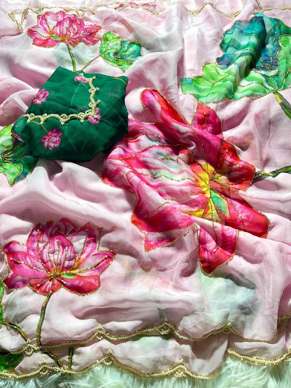 A close-up of a light pink embroidered fabric with large lotus flowers in pink, green, and red, featuring gold zari embroidery and a scalloped border. A folded dark green embroidered fabric piece is placed on top.