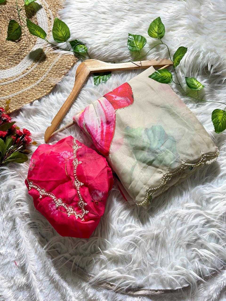 A folded pink and green floral printed fabric accessory with scalloped edges, displayed on a wooden hanger next to a bright pink embroidered fabric piece on a white faux fur surface with decorative green leaves.