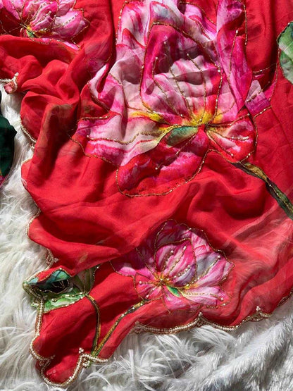 A close-up of a red sheer fabric with large pink and white floral prints, gold embroidered outlines, and green leaf accents, draped over a white faux fur surface.