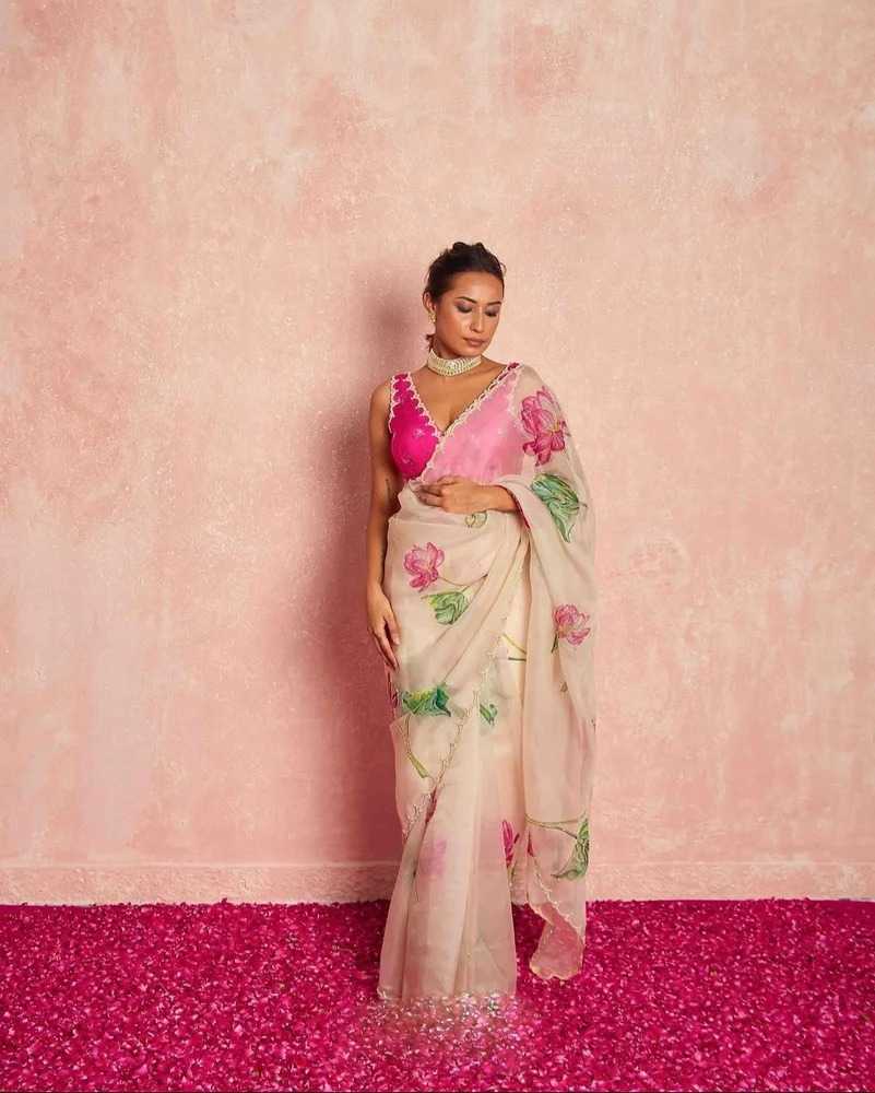 A woman models a traditional Indian saree with a vibrant pink blouse and a cream-colored saree featuring pink floral prints, standing against a textured pink wall and a bright pink floor.