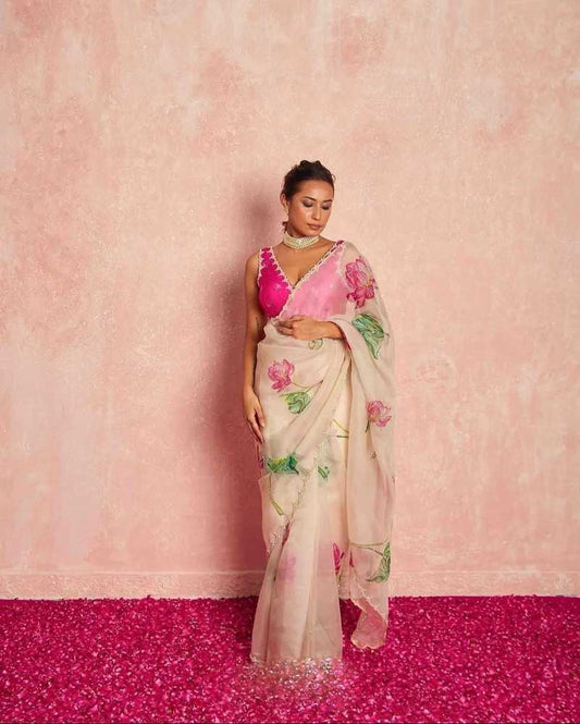 A woman models a traditional Indian saree with a vibrant pink blouse and a cream-colored saree featuring pink floral prints, standing against a textured pink wall and a bright pink floor.