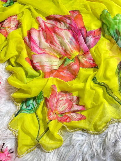 A bright yellow sheer fabric scarf or dupatta featuring large pink and coral floral prints with green leaves and a scalloped gold-trimmed edge, laid on a white textured surface.