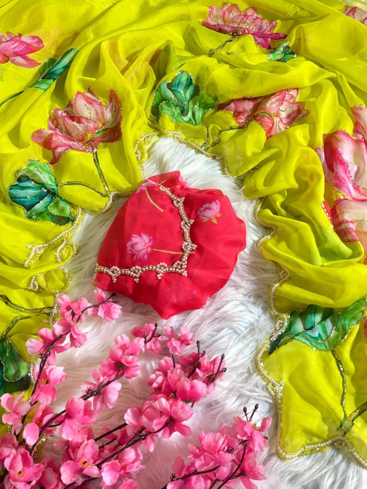 A bright yellow embroidered dupatta with pink lotus flowers and green leaves, styled with a red embroidered blouse piece and pink artificial blossoms on a white faux fur background.