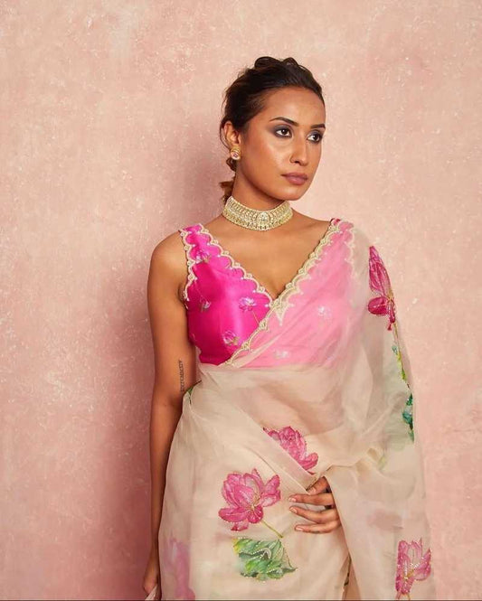 A woman models a traditional Indian saree with a vibrant pink embroidered blouse and a sheer cream-colored drape decorated with pink lotus motifs, set against a textured pink background.