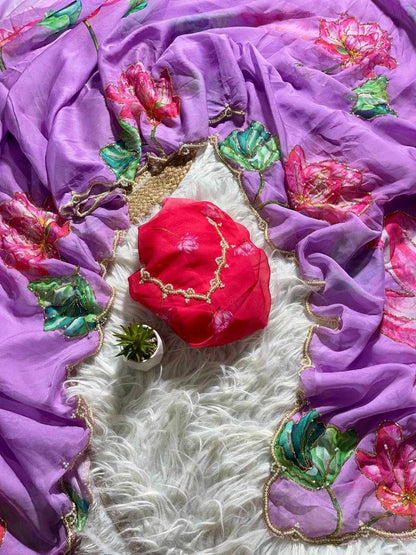 A purple embroidered dupatta or scarf laid out on a white faux-fur surface, featuring pink floral motifs and green leaf embroidery with gold borders. A bright red fabric pouch with gold trim rests in the center, alongside a small succulent.