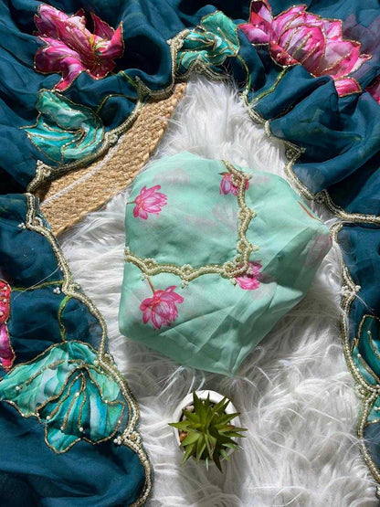 A mint green embroidered fabric pouch or small bag with pink floral motifs and gold lace trim, styled on a white faux-fur surface with a teal embroidered textile and a small succulent around it.