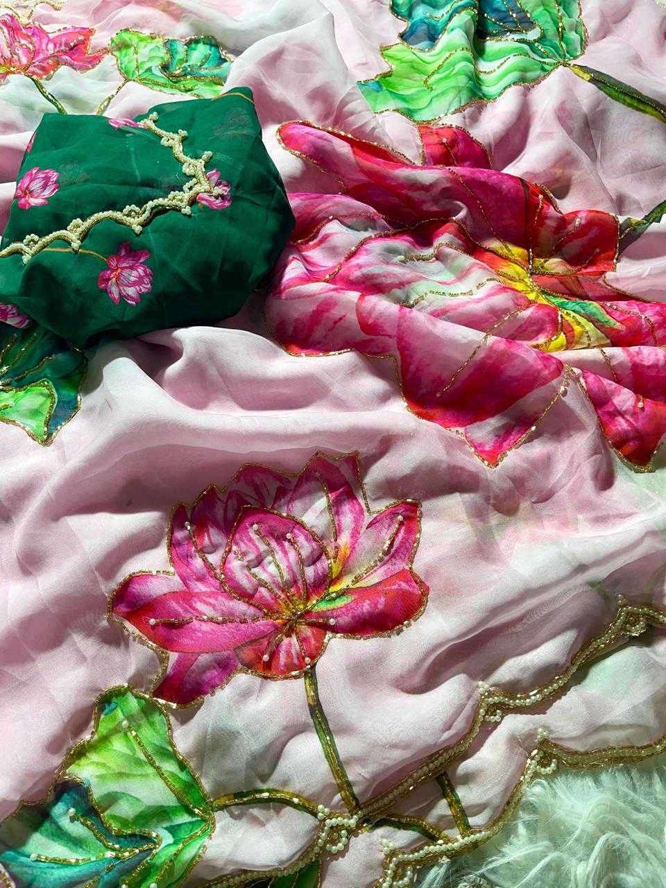 A close-up of a pink and white floral fabric with embroidered lotus flowers in pink, green, and yellow, featuring gold zari borders and a dark green fabric piece with pink floral embroidery.