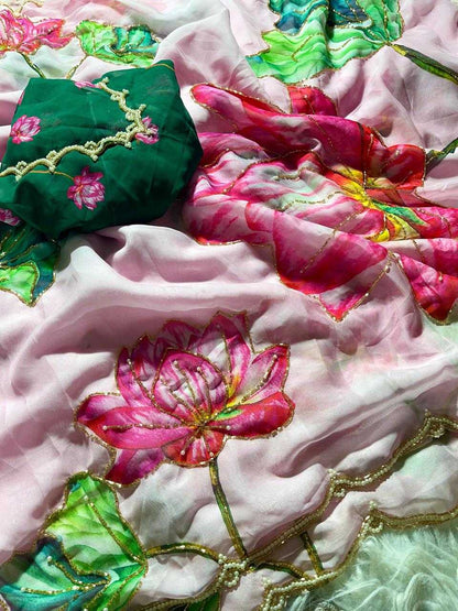 A close-up of a pink and white floral fabric with embroidered lotus flowers in pink, green, and yellow, featuring gold zari borders and a dark green fabric piece with pink floral embroidery.