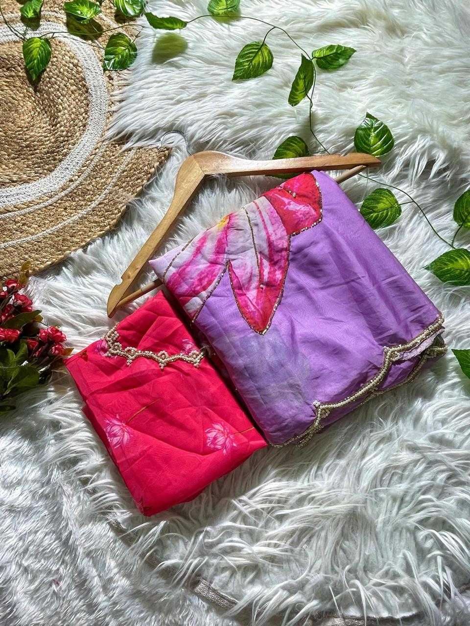 Two folded fabric garments or textiles displayed on a wooden hanger over a white faux-fur surface, with decorative green leaves, a woven hat, and small red flowers around them. One item is bright pink/coral and the other is lavender-purple with a floral print in pink, red, and white, both with gold trim.
