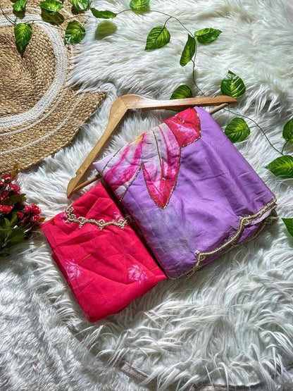 Two folded fabric garments or textiles displayed on a wooden hanger over a white faux-fur surface, with decorative green leaves, a woven hat, and small red flowers around them. One item is bright pink/coral and the other is lavender-purple with a floral print in pink, red, and white, both with gold trim.