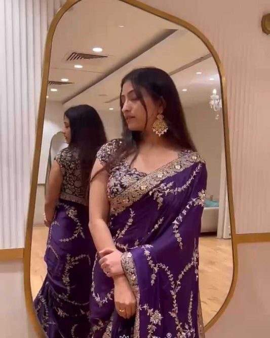 Woman in a purple saree with gold details standing in front of a mirror.