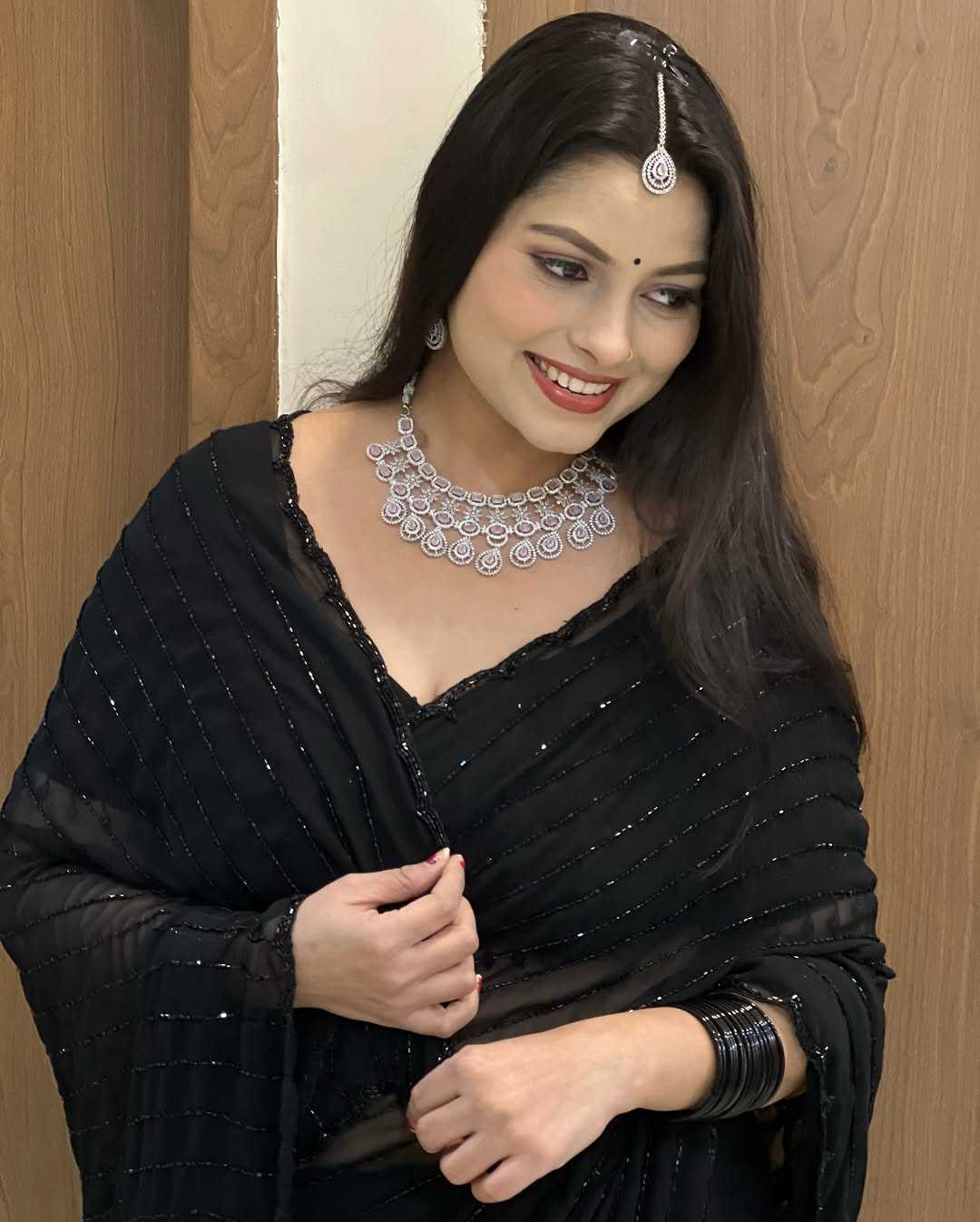 Woman wearing a black saree with silver jewelry against a wooden door background