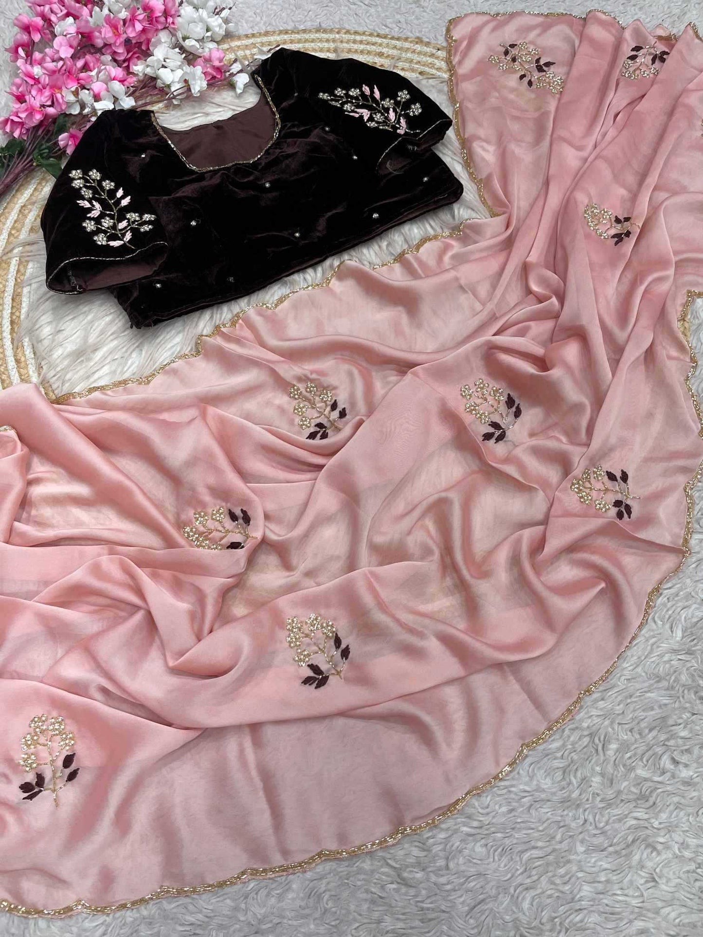 A pink draped fabric garment with scattered floral embroidery and a scalloped gold-trimmed edge, shown alongside a black embroidered blouse on a white textured surface with decorative flowers in the corner.