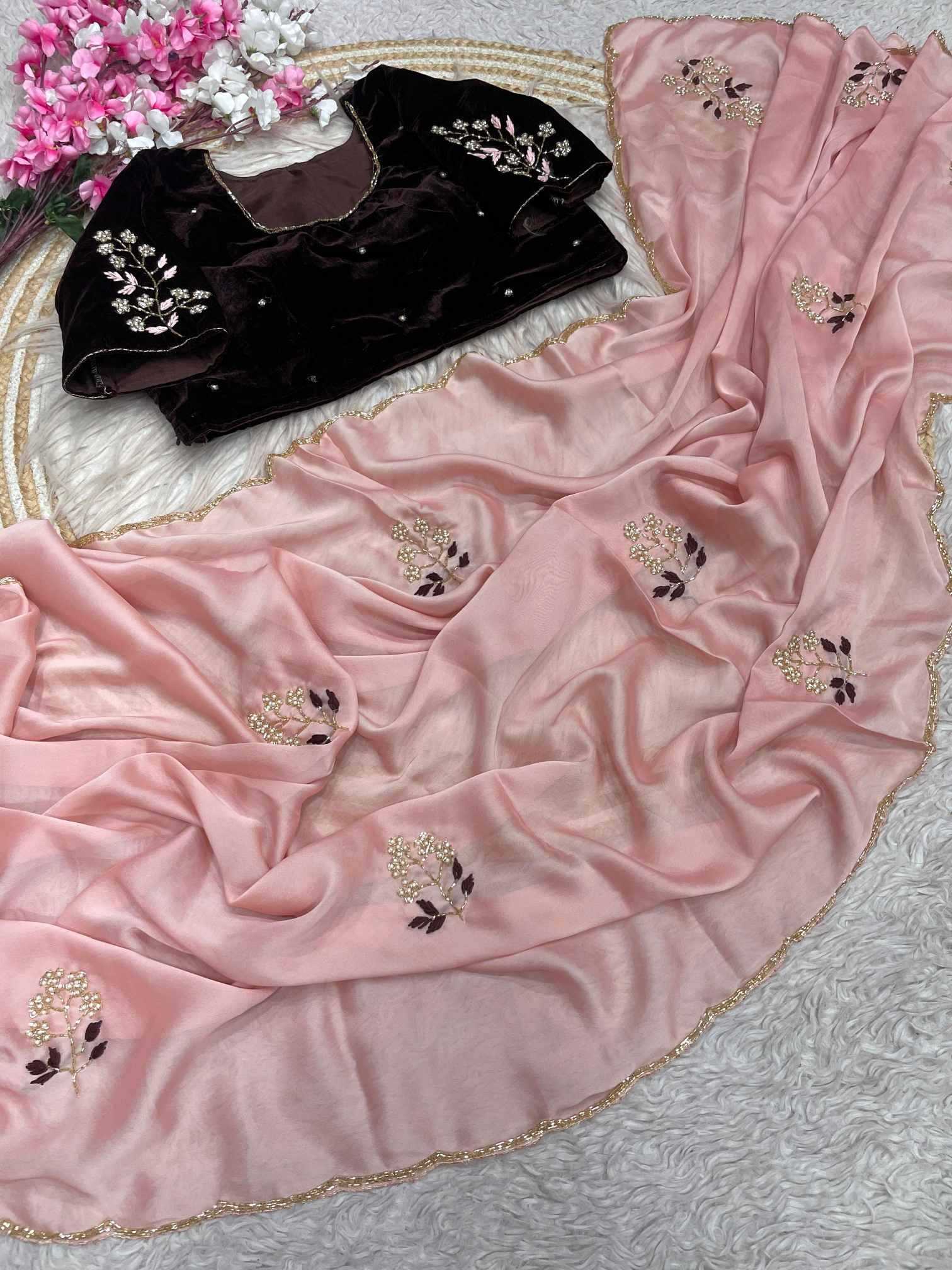 A pink draped fabric garment with scattered floral embroidery and a scalloped gold-trimmed edge, shown alongside a black embroidered blouse on a white textured surface with decorative flowers in the corner.