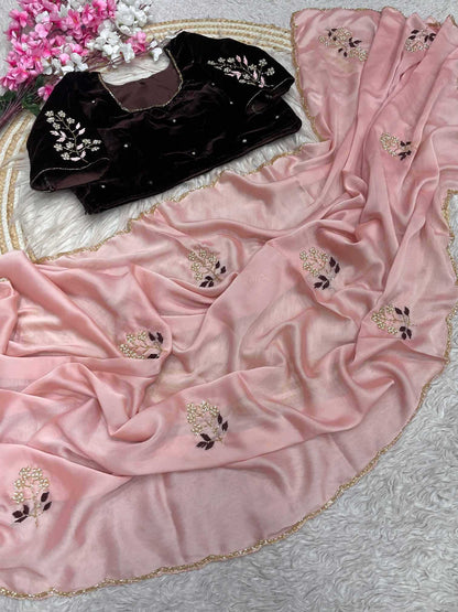 A pink draped fabric garment with scattered floral embroidery and a scalloped gold-trimmed edge, shown alongside a black embroidered blouse on a white textured surface with decorative flowers in the corner.