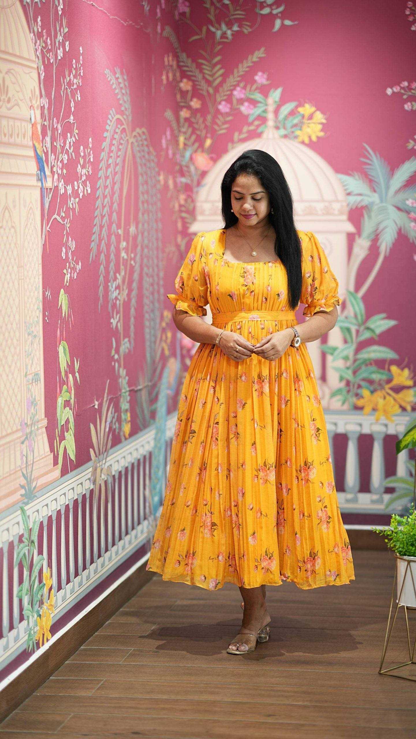 Woman in a yellow floral dress standing in front of a decorative wall with pink and white patterns.