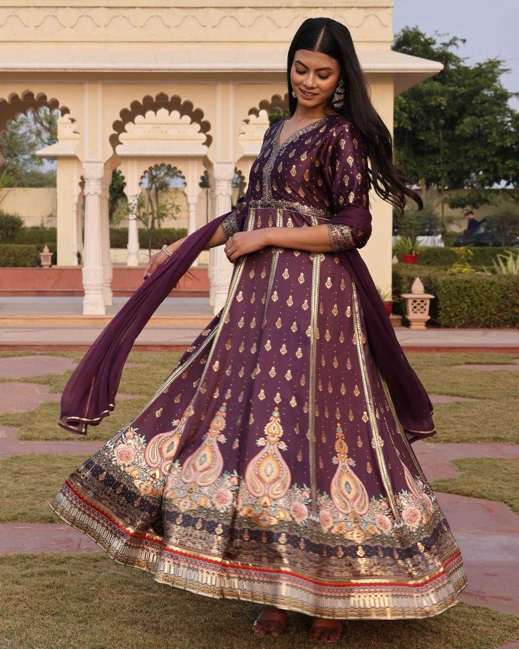 Woman in a traditional purple embroidered dress standing outdoors with a decorative archway in the background.