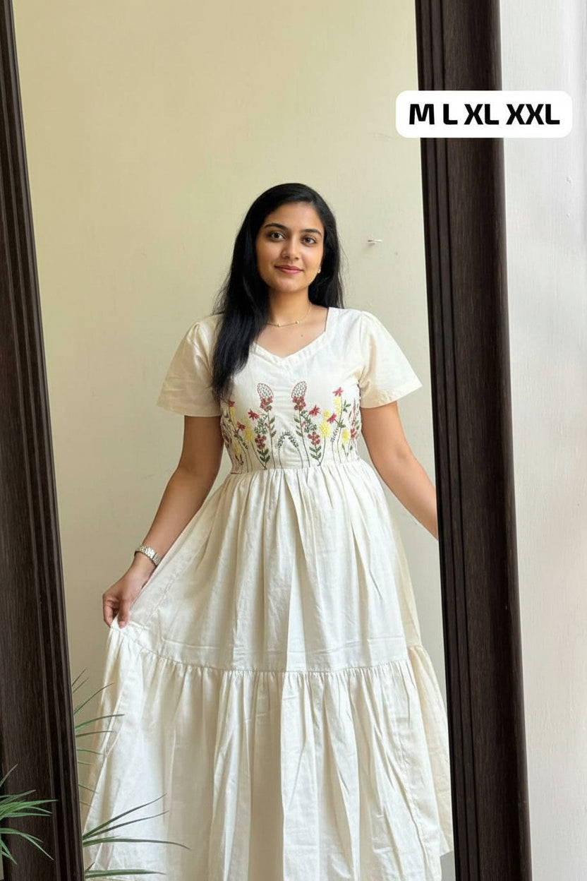 A woman takes a mirror selfie wearing a cream-colored tiered maxi dress with short sleeves and a floral embroidered bodice. The dress has a smocked waist and is shown in an indoor setting with a size overlay label reading 'M L XL XXL'.