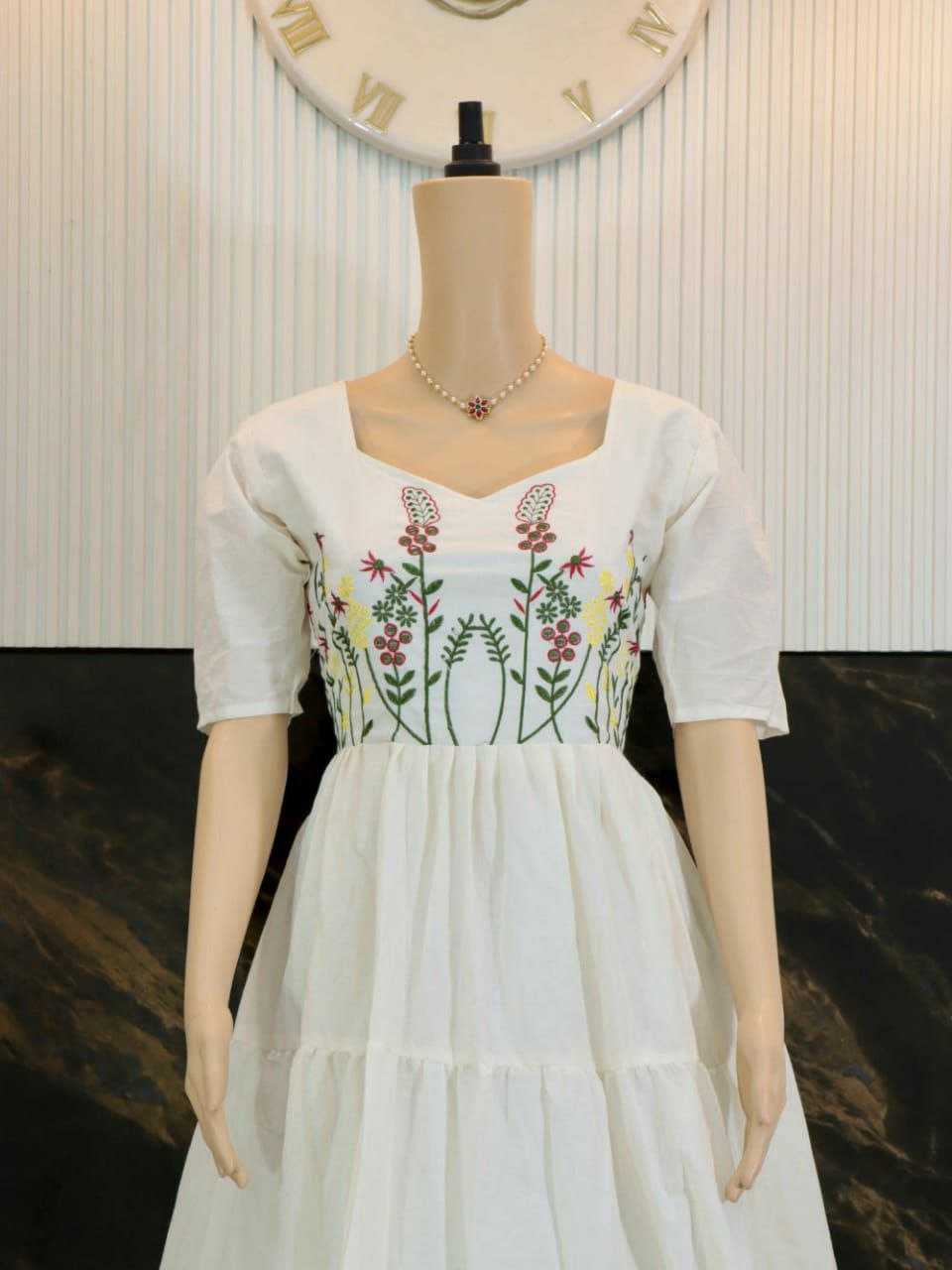 A white tiered dress displayed on a mannequin, featuring short puff sleeves, a square neckline, and colorful floral embroidery across the bodice.
