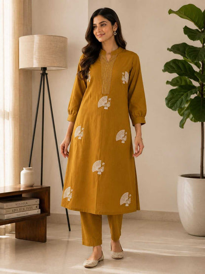 Woman wearing a mustard yellow kurta with white patterns in a room with a lamp and plant.