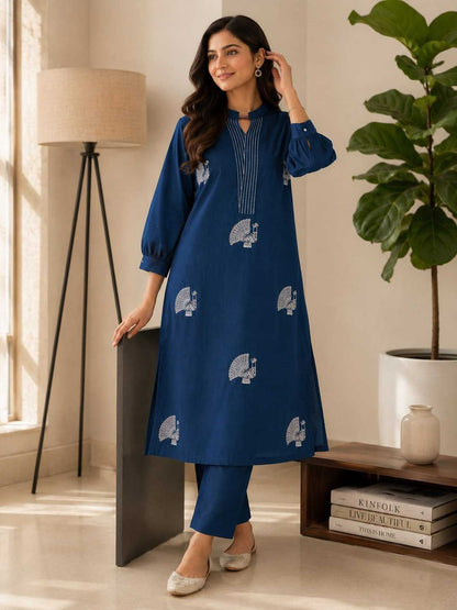 Woman in a blue dress with white patterns standing in a room with a lamp and plant.