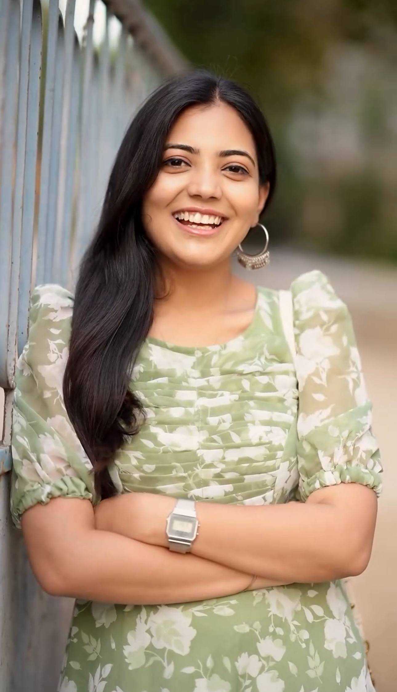 A woman with long black hair smiling, wearing a light green and white floral print short-sleeve top, standing outdoors with her arms crossed.