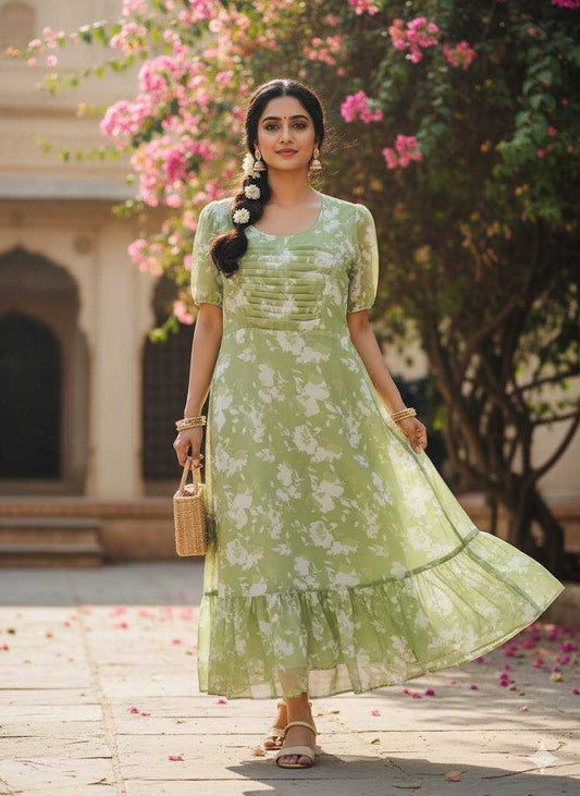 A woman wearing a light green and white floral print midi dress with short sleeves and a tiered ruffled hem, standing outdoors in front of a building with pink flowering trees.