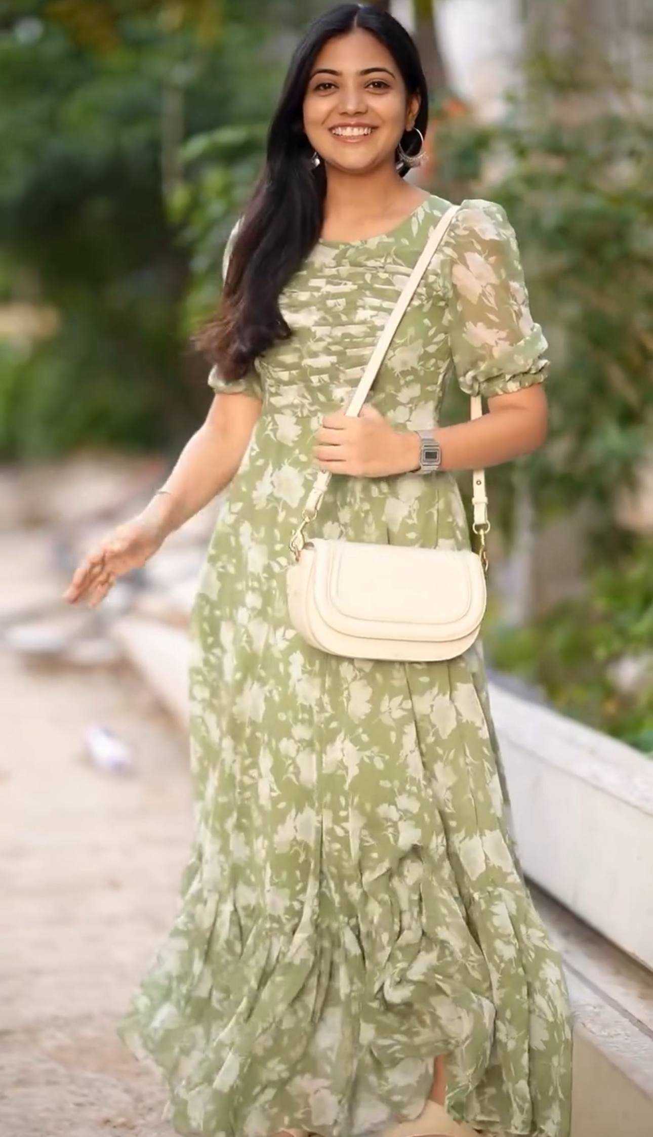 A woman wearing a light green and white tie-dye maxi dress with puff sleeves, carrying a cream-colored crossbody bag, standing outdoors on a paved path with green foliage in the background.