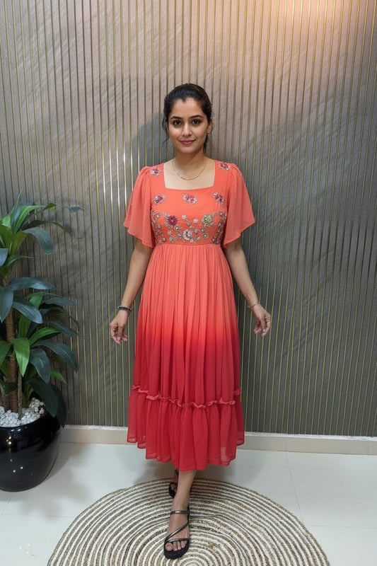 A woman takes a mirror selfie wearing a short-sleeved tiered maxi dress with a red-to-orange ombre gradient and floral embroidery on the bodice. She is standing on a round woven rug in front of a slatted wood wall.