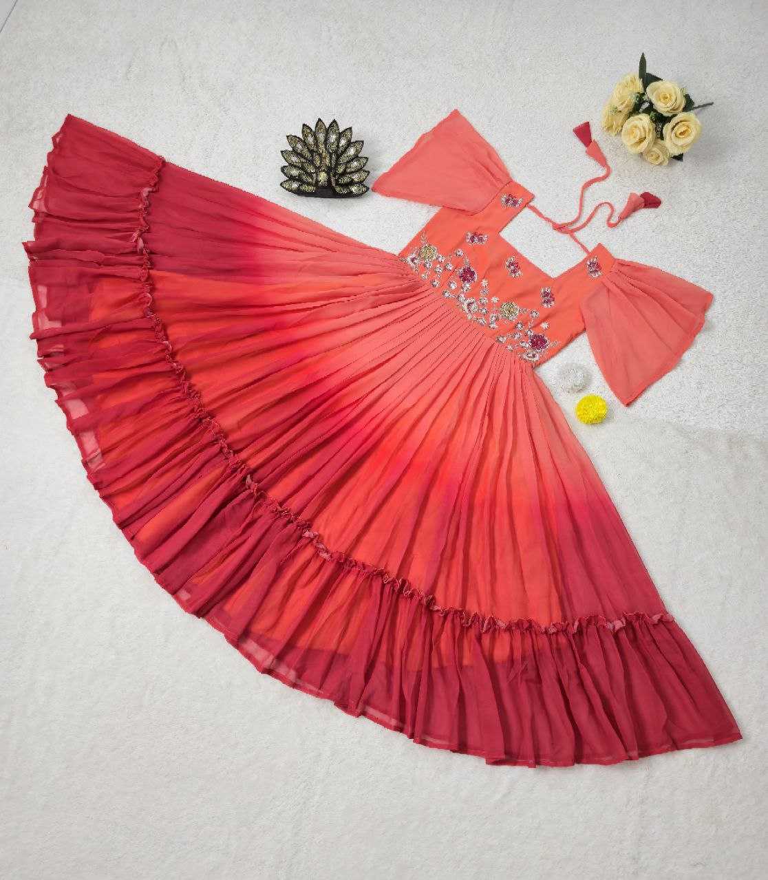A red and orange gradient baby dress with tiered ruffles, laid flat on a white textured background alongside decorative props including a small flower arrangement, a metallic ornament, and a yellow pom-pom.