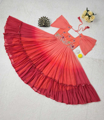 A red and orange gradient baby dress with tiered ruffles, laid flat on a white textured background alongside decorative props including a small flower arrangement, a metallic ornament, and a yellow pom-pom.