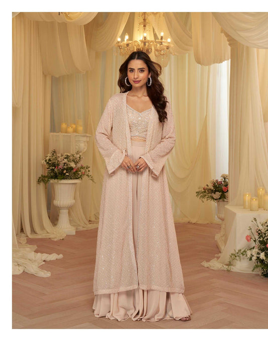 Woman in a beige traditional outfit standing in a decorated room with floral arrangements and candles.