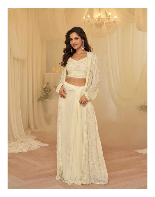 A woman modeling a cream-colored traditional Indian outfit consisting of a long-sleeved crop top, a matching skirt, and a sheer embroidered outer layer, set against a warm beige draped backdrop with a chandelier.